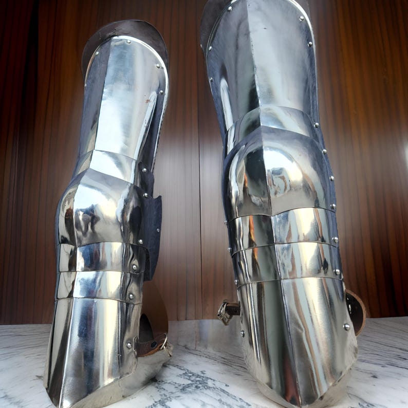 May include: A pair of polished steel greaves designed to protect the lower legs. These metallic armor pieces feature articulated plates, visible rivets, and brown leather straps with metal buckles for secure fastening around the calf area.