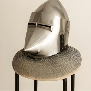 May include: A polished silver knight's helmet with a pointed top and a long, angled face guard. The helmet rests on a circular, chainmail-covered cushion atop a black metal stool. The helmet has small eye slits and ventilation holes.