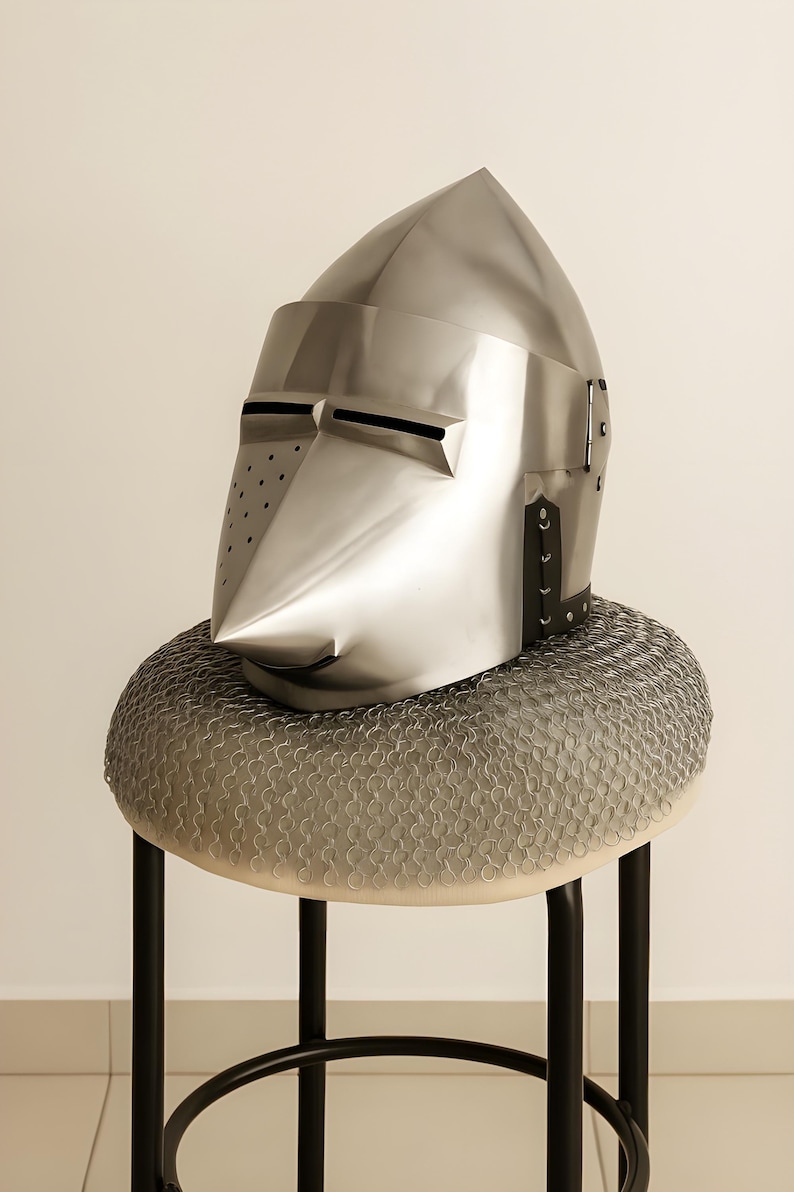 May include: A polished silver knight's helmet with a pointed top and a long, angled face guard. The helmet rests on a circular, chainmail-covered cushion atop a black metal stool. The helmet has small eye slits and ventilation holes.