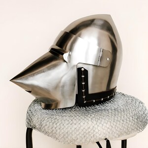 May include: A polished silver medieval helmet with a pointed visor and a hinged visor plate. The helmet rests on a chainmail coif, which is placed on a small stool. The helmet is a historical item.