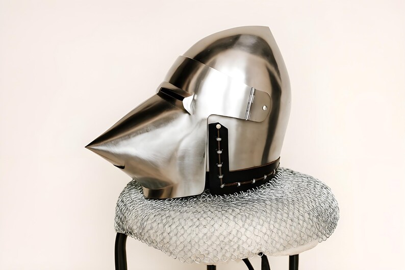 May include: A polished silver medieval helmet with a pointed visor and a hinged visor plate. The helmet rests on a chainmail coif, which is placed on a small stool. The helmet is a historical item.