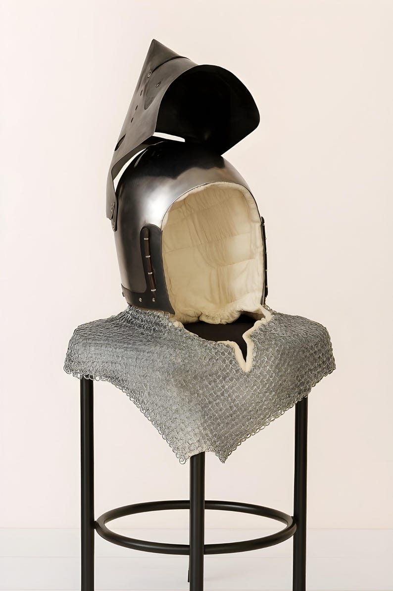 May include: A medieval helmet with a hinged visor and a chainmail neck guard. The helmet is black with a padded interior. The chainmail is silver. The helmet is displayed on a black stand.