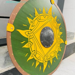 Op de afbeelding: Een rond houten schild met een groen midden met een geel zonontwerp. Het schild heeft een bruine rand met messing klinknagels en gele hoekaccenten. In het midden zit een zwarte metalen baas.