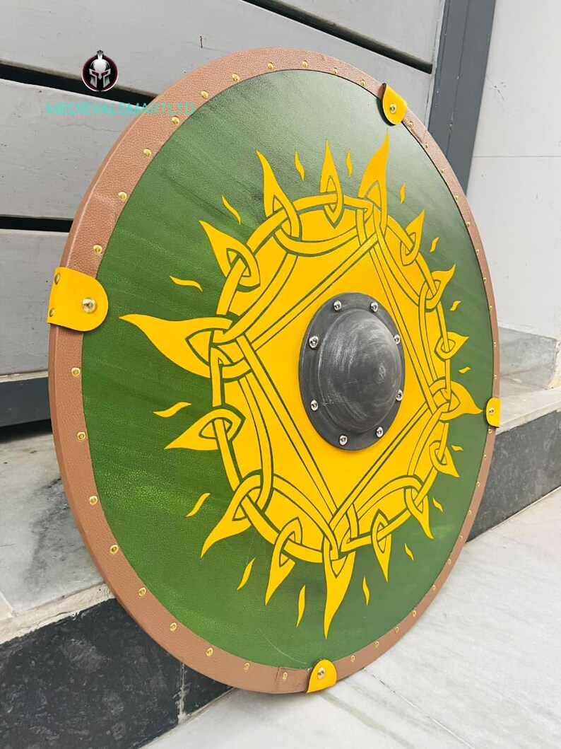 Op de afbeelding: Een rond houten schild met een groen midden met een geel zonontwerp. Het schild heeft een bruine rand met messing klinknagels en gele hoekaccenten. In het midden zit een zwarte metalen baas.