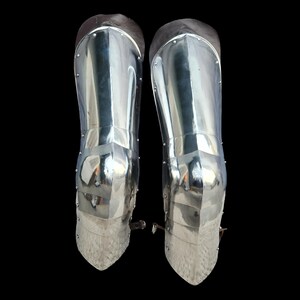 May include: A pair of polished silver-colored metal greaves for lower leg protection. These articulated armor pieces feature a smooth, reflective surface with visible rivets along the sides and brown leather straps for secure fastening.