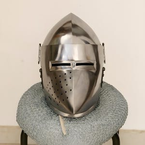 May include: A polished, silver-colored medieval helmet with a pointed top and a visor featuring horizontal eye slits. The helmet has small ventilation holes and is displayed on a chainmail coif.