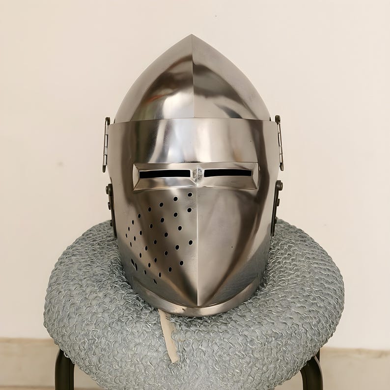 May include: A polished, silver-colored medieval helmet with a pointed top and a visor featuring horizontal eye slits. The helmet has small ventilation holes and is displayed on a chainmail coif.