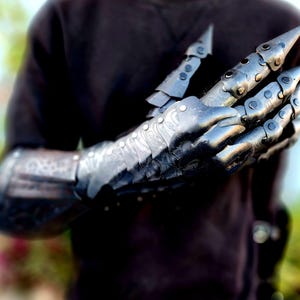 May include: A black metal gauntlet with articulated fingers and sharp claws. The gauntlet is worn on a person's arm, and the fingers are curled into a fist.