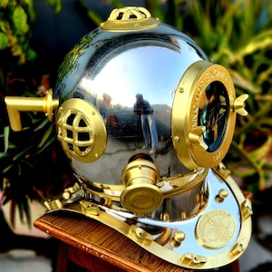 Op de afbeelding: Een zilveren en gouden duikhelm met een messing kijkvenster en de tekst "Anchor Engine" aan de zijkant. De helm staat op een houten standaard.