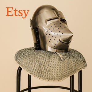 May include: A polished metal knight's helmet with a pointed visor and numerous small ventilation holes. The helmet rests on a chainmail coif, displayed on a black metal stand. The word "Etsy" is in orange text in the upper left corner.