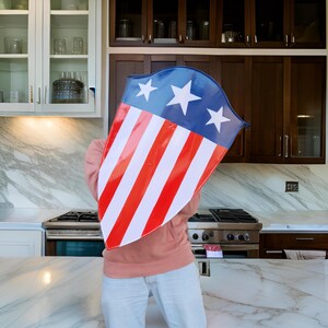 Puede incluir: Una persona sosteniendo un escudo rojo, blanco y azul con estrellas. El escudo est&aacute; cubriendo la cara de la persona. La persona est&aacute; de pie en una cocina con gabinetes blancos y una encimera de m&aacute;rmol.