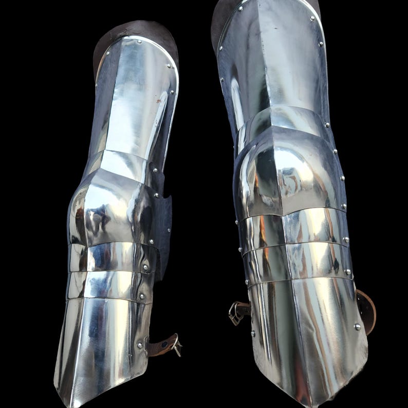 May include: A pair of polished steel greaves designed to protect the lower legs. These metallic armor pieces feature articulated knee sections, multiple rivets along the side seams, and brown leather straps with metal buckles for secure fastening.