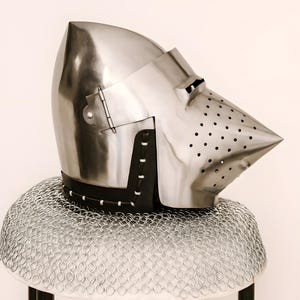 May include: A polished steel helmet with a pointed visor and a series of small holes for ventilation. The helmet is set on a chainmail coif and a black leather neck guard. The helmet is a historical item.