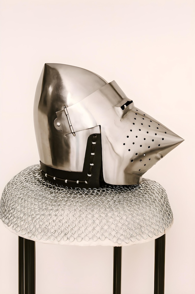 May include: A polished steel helmet with a pointed visor and a series of small holes for ventilation. The helmet is set on a chainmail coif and a black leather neck guard. The helmet is a historical item.