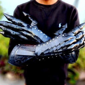May include: A pair of black metal gauntlets with spiked knuckles and articulated fingers. The gauntlets are worn by a person, but the focus is on the gauntlets themselves.