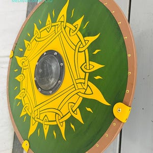 Op de afbeelding: Een rond schild met een groen midden en een gele zonnestraal. Het schild heeft een bruine rand met messing klinknagels en gele leren riemen. Het midden heeft een zwarte metalen schildknop. De tekst "MEDIEVALSMITHS LTD" is zichtbaar.