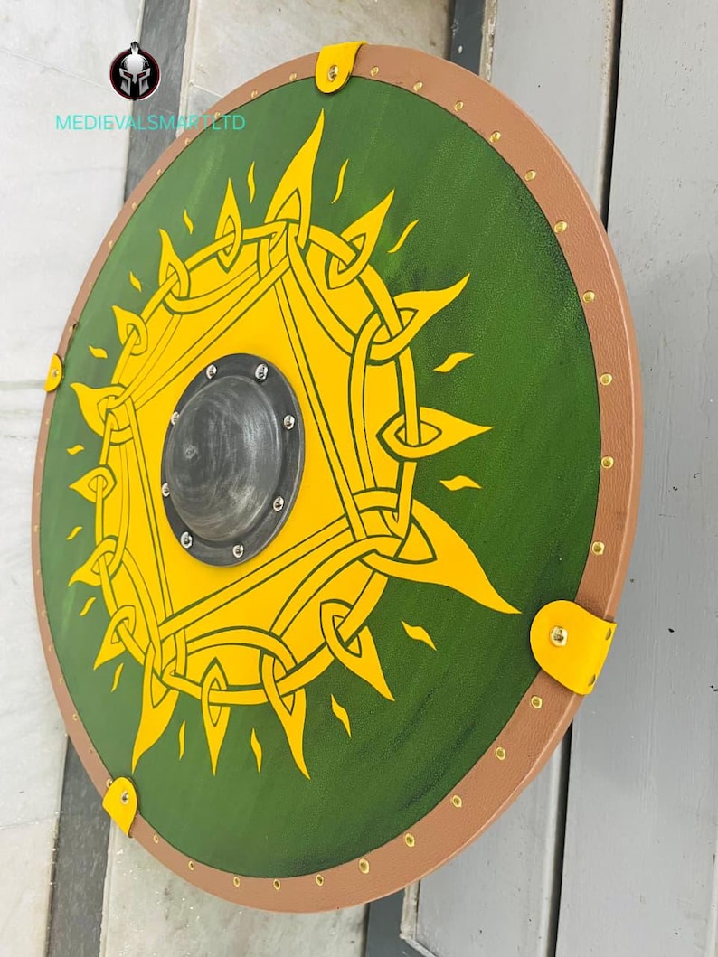 Op de afbeelding: Een rond schild met een groen midden en een gele zonnestraal. Het schild heeft een bruine rand met messing klinknagels en gele leren riemen. Het midden heeft een zwarte metalen schildknop. De tekst "MEDIEVALSMITHS LTD" is zichtbaar.