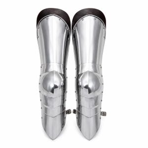 May include: A pair of polished silver-colored metal greaves featuring articulated plates, a central knee cap, and dark brown leather trim at the top. Each piece is constructed with visible rivets along the sides and includes a small metal buckle strap.