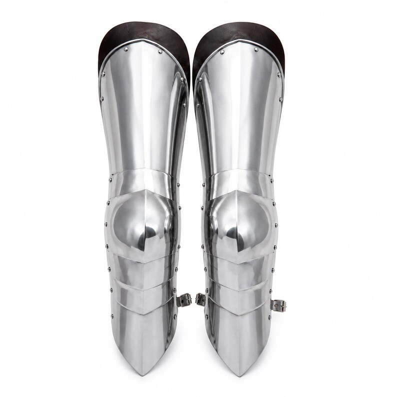 May include: A pair of polished silver-colored metal greaves featuring articulated plates, a central knee cap, and dark brown leather trim at the top. Each piece is constructed with visible rivets along the sides and includes a small metal buckle strap.
