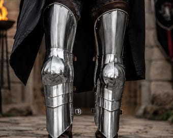 May include: Polished steel leg armor featuring articulated greaves and poleyns with visible rivets and leather straps. The metal plating covers the shins and knees, worn over dark quilted fabric trousers and brown leather boots with metal buckles.