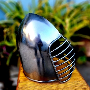 Puede incluir: Un casco plateado con una protección facial de malla de alambre. El casco está hecho de metal y tiene un acabado liso y brillante.