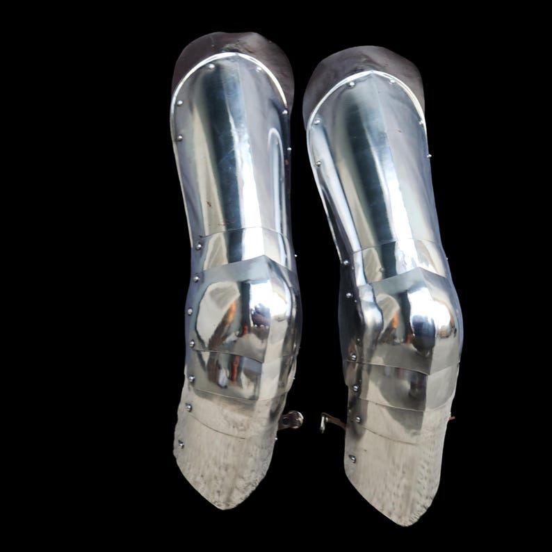May include: A pair of polished steel greaves and poleyns designed for leg protection. These articulated metal armor pieces feature a smooth, reflective surface with visible rivets along the sides and leather straps for secure fastening.
