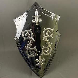 May include: A decorative shield with a black and silver design. The shield has a glossy black centre with silver trim and ornate silver embellishments. The edges are lined with small silver rivets and a repeating triangle pattern.