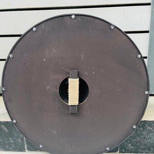Op de afbeelding: Een ronde, donkerbruine schild met een zwarte rand en zilveren klinknagels. De achterkant heeft een centrale handgreep omwikkeld met natuurlijk touw en een rechthoekige houten steun. Geschikt voor historische re-enactment of display.
