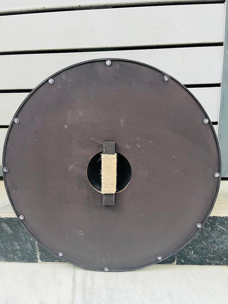 Op de afbeelding: Een ronde, donkerbruine schild met een zwarte rand en zilveren klinknagels. De achterkant heeft een centrale handgreep omwikkeld met natuurlijk touw en een rechthoekige houten steun. Geschikt voor historische re-enactment of display.