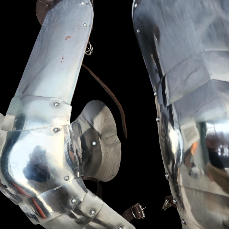 May include: A pair of polished steel leg armor pieces featuring articulated knee plates, curved side wings, and metal rivets. The armor includes brown leather straps with metal buckles for fastening, showing a reflective metallic surface finish.