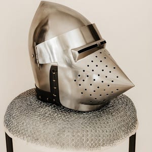 May include: A polished metal knight's helmet with a pointed visor and numerous small ventilation holes. The helmet rests on a chainmail coif atop a small stool with black legs. The helmet's design is reminiscent of medieval armor.