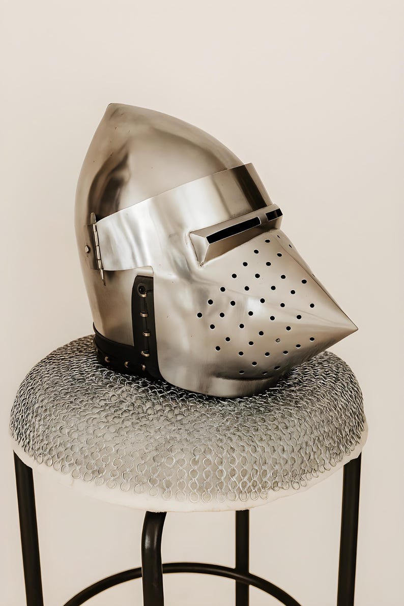 May include: A polished metal knight's helmet with a pointed visor and numerous small ventilation holes. The helmet rests on a chainmail coif atop a small stool with black legs. The helmet's design is reminiscent of medieval armor.