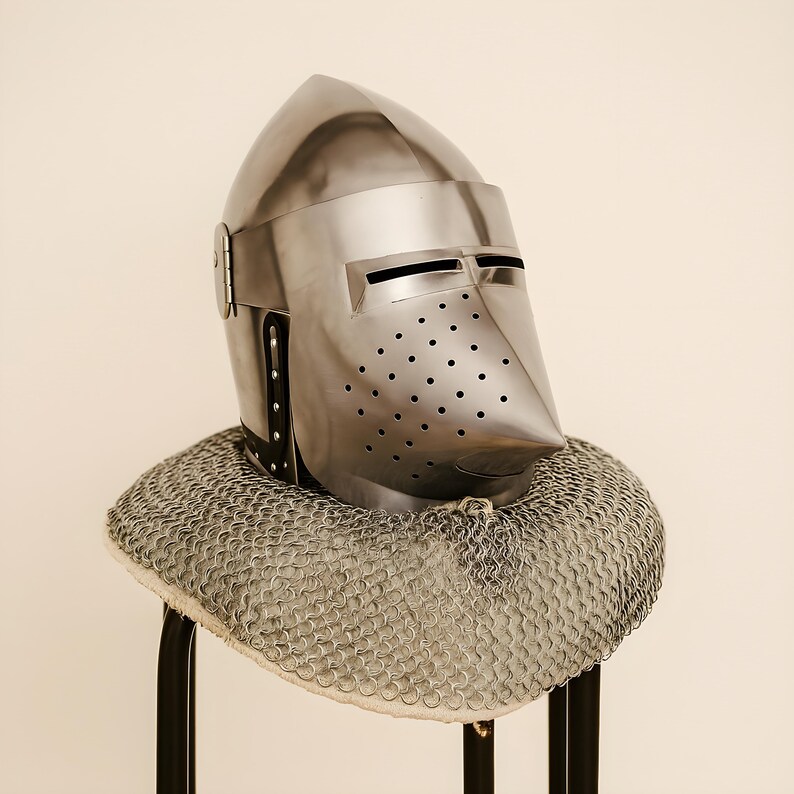 May include: A polished steel knight's helmet with a pointed visor and numerous small ventilation holes. The helmet rests on a chainmail coif, showcasing medieval armor design. The helmet is displayed on a black stand.