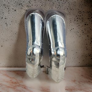 May include: A pair of polished silver-colored metal greaves designed to protect the lower legs. These pieces of armor feature a smooth, reflective surface with visible rivets along the edges and a segmented construction that tapers toward the ankle.
