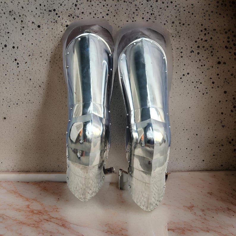 May include: A pair of polished silver-colored metal greaves designed to protect the lower legs. These pieces of armor feature a smooth, reflective surface with visible rivets along the edges and a segmented construction that tapers toward the ankle.
