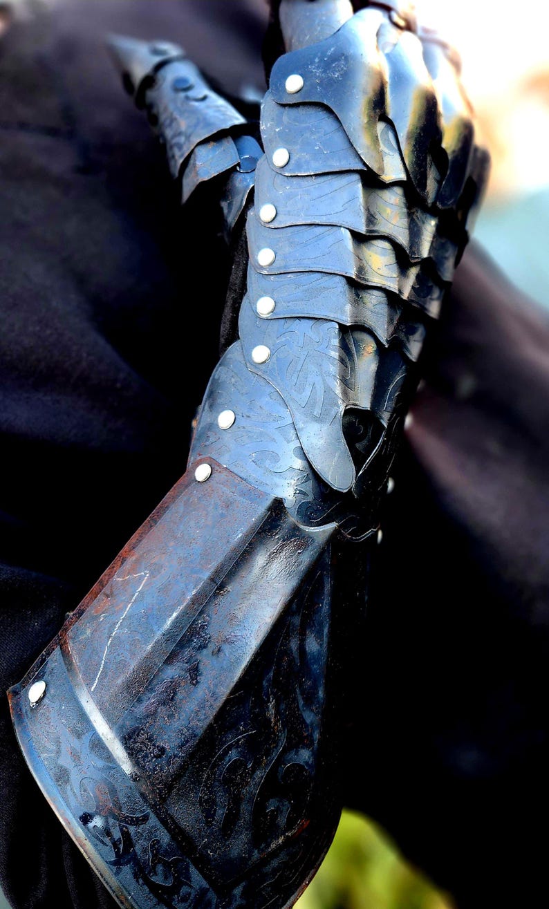 May include: A black metal gauntlet with decorative details and rivets. The gauntlet is worn on a person's left hand and extends to the elbow.