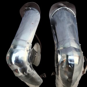 May include: A pair of polished steel leg armor pieces featuring articulated knee cops and thigh plates. The metal segments are connected by rivets and include brown leather straps with metal buckles for secure attachment to the wearer's legs.