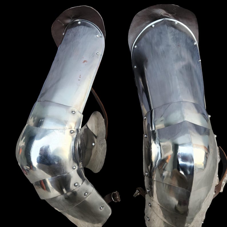 May include: A pair of polished steel leg armor pieces featuring articulated knee cops and thigh plates. The metal segments are connected by rivets and include brown leather straps with metal buckles for secure attachment to the wearer's legs.