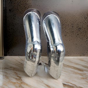 May include: A pair of polished silver-colored metal greaves designed to protect the lower legs. These articulated armor pieces feature a smooth, reflective surface with visible rivets along the sides, constructed in multiple overlapping segments.