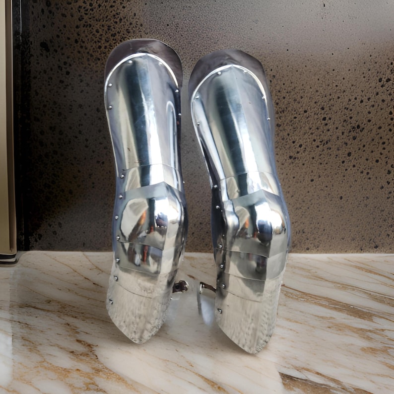 May include: A pair of polished silver-colored metal greaves designed to protect the lower legs. These articulated armor pieces feature a smooth, reflective surface with visible rivets along the sides, constructed in multiple overlapping segments.