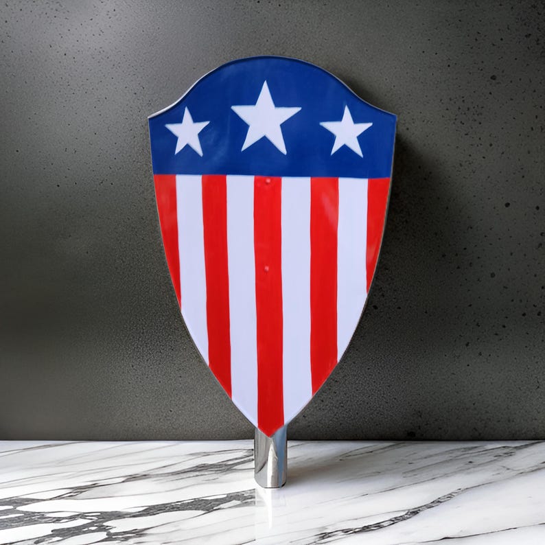 Puede incluir: Un tap&oacute;n de botella en forma de escudo rojo, blanco y azul con una base plateada. El escudo tiene cinco rayas rojas y blancas y tres estrellas blancas sobre un fondo azul.