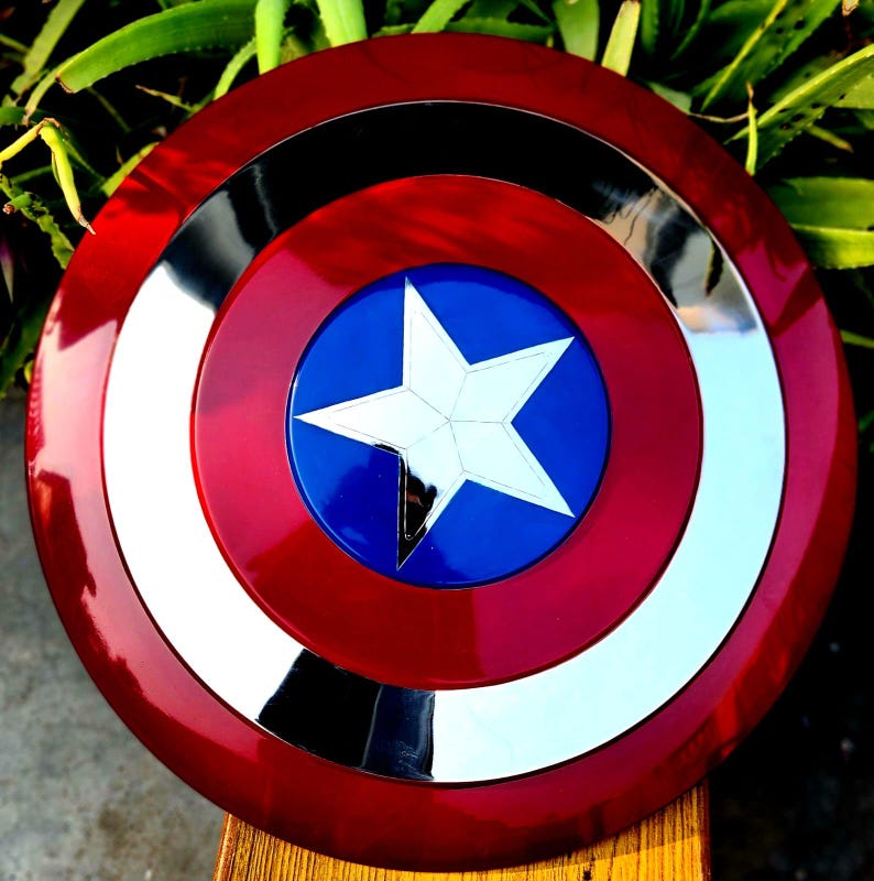 May include: A red, white and blue Captain America shield with a silver star in the centre. The shield is made of metal and has a shiny finish.