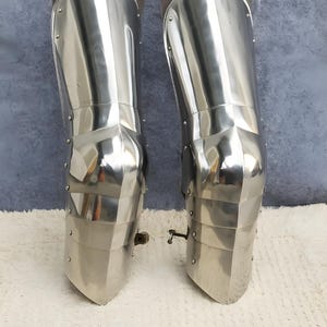 May include: A pair of polished steel leg armor greaves featuring articulated knee sections and metal rivets. The armor pieces are constructed with smooth, reflective surfaces and include brown leather straps at the top for secure attachment.