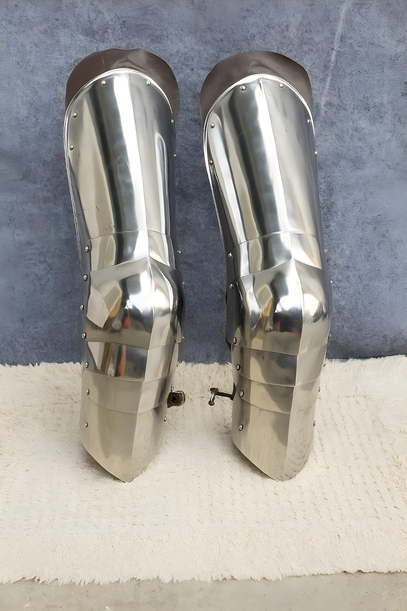 May include: A pair of polished steel leg armor greaves featuring articulated knee sections and metal rivets. The armor pieces are constructed with smooth, reflective surfaces and include brown leather straps at the top for secure attachment.