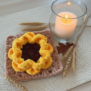 Peut inclure: Un dessous de verre en forme de tournesol au crochet, avec un centre marron et des pétales jaunes, posé sur un dessous de verre carré rose. Une bougie allumée dans un verre et des épis de blé. Décoration intérieure.