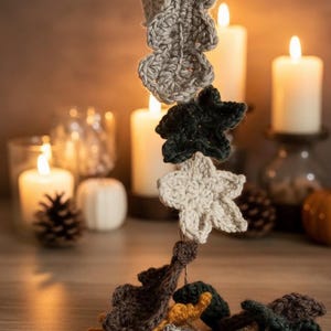 May include: A handmade crochet leaf garland in various fall colors, including mustard yellow, gray, and dark green. The leaves are suspended against a warm, candlelit background, creating a cozy autumn aesthetic.