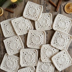 May include: A collection of cream-coloured, square crochet coasters arranged on a wooden surface. Each coaster features a circular, textured design in the centre, surrounded by a square border. The image includes decorative elements like cinnamon sticks and dried orange slices.