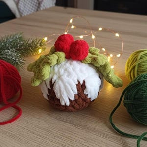 以下が含まれることがあります： かぎ針編みのクリスマスプディングオーナメント。オーナメントは茶色、白、緑色で、赤いベリーのアクセントが付いています。プディングは、ストリングライトと毛糸玉のある木製の表面にあります。