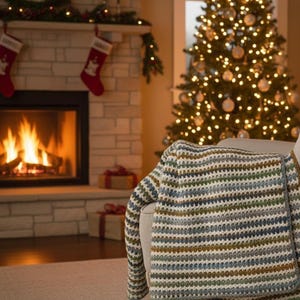 Può includere: Una coperta a righe blu, verdi e marroni è drappeggiata su una poltrona. La scena comprende un camino acceso, calze di Natale e un albero di Natale decorato, creando un'atmosfera calda e festosa.