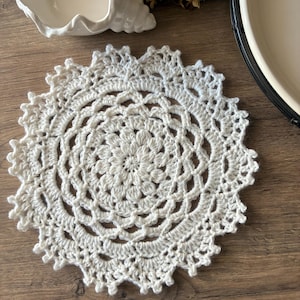 May include: A cream-coloured, crocheted doily with an intricate circular pattern. The doily sits on a wooden surface, highlighting its detailed design and texture. This handmade piece adds a touch of vintage charm to any home decor.